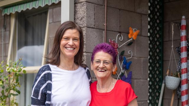 Two women side by side smiling at the camera and standing in front of a house