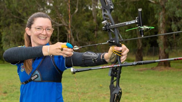 Woman in blue shirt holds archery bow and arrow.