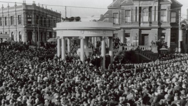 Black and white historical photo shows circular memorial surrounded by crowds of people