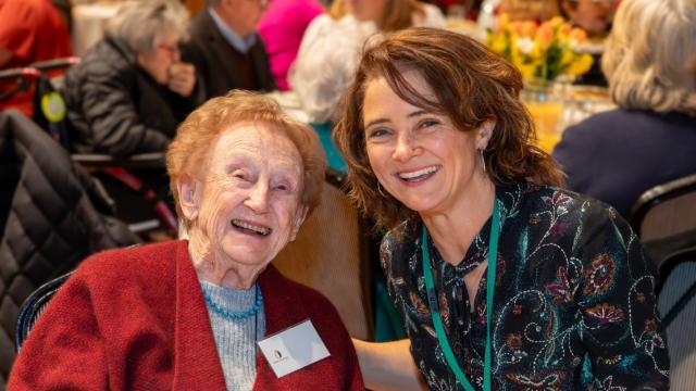 A senior citizen with a younger person seated in a room with banquet tables