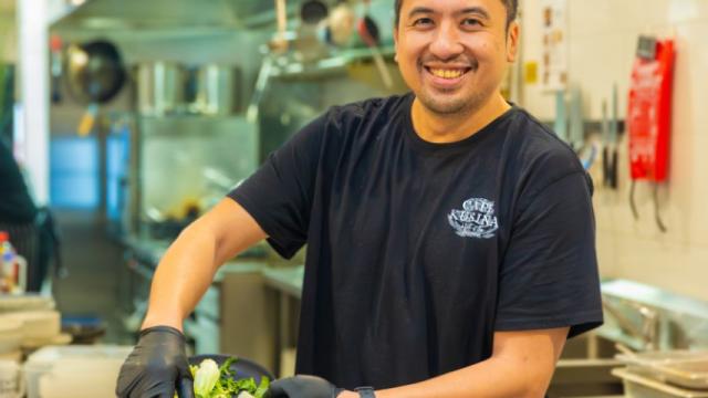 Man stands smiling in café kitchen while emptying vegetable scraps into container.