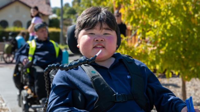 Three people in wheelchairs on path in playground, with boy smiling at front and house and trees in background.