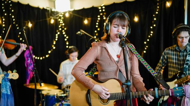 A 4-piece band play musical instruments. Decorative fairylights can be seen in the background. 