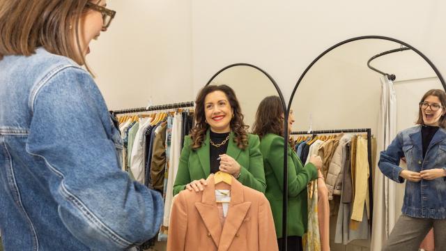 2 women in a clothing store, one is the sales assistant holding up a jacket while the other is a customer trying on a jacket.