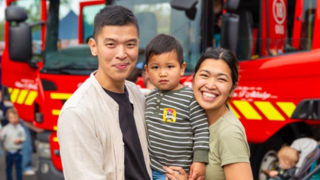 Smiling man and woman stand holding small child with fire engine in background