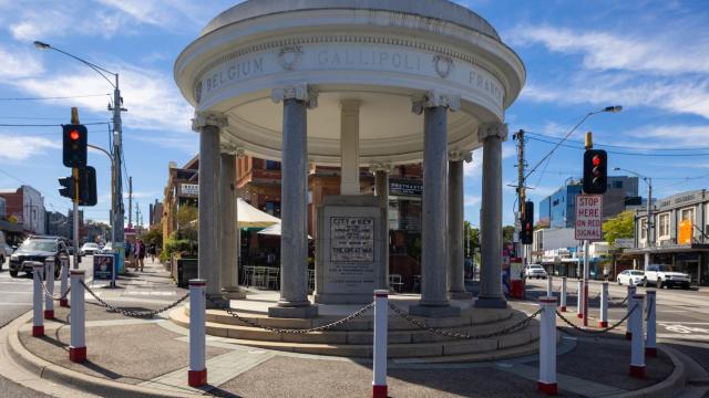 Street view of a cenotaph building on the edge of a roundabout with a historic building in background