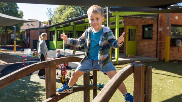 Preschool child stands on wooden bridge in playground giving thumbs-up