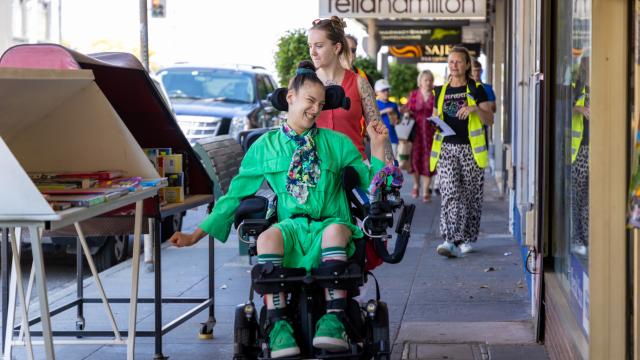 Young woman in wheelchair moves down footpath beside shops and display stands.