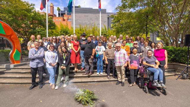 A group of people of diverse nationalities, genders and abilities, gather under the flag poles at Hawthorn Arts Centre. There is the progress pride flag, Aboriginal flag and the Torres Straight Islander flag.