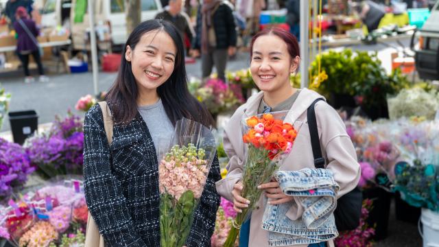 2 people standing in front of a flower stall holding bunches of flowers and smiling.