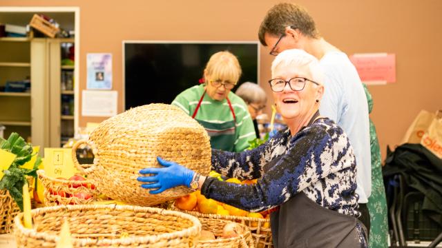 Smiling person in apron holds basket over table holding fresh fruit and vegetables