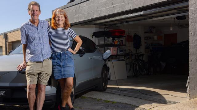 Two people stand smiling in front of grey car outside garage