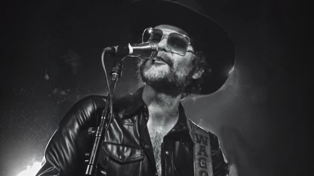 Black and white photo of man wearing a leather jacket, sunglasses and a cowboy hat standing at a microphone playing the guitar.