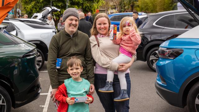 Two adults with 2 children stand smiling between rows of electric vehicles