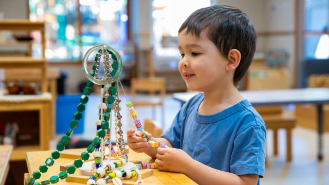 A jewellery stand is on a table in front of a child. The child is practicing threading beads onto string.