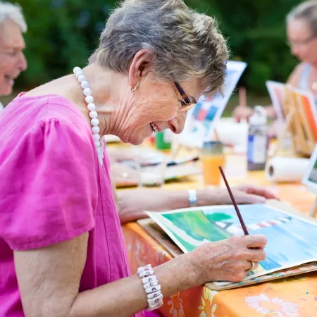An older woman wearing a pink top paints on a table