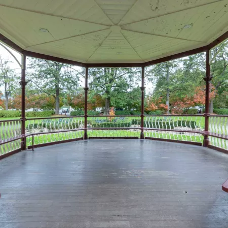 Inside view of round outdoor rotunda located in grass area, with trees in the distance. The columns and bench seats are dark red and the roof and balustrades are white.