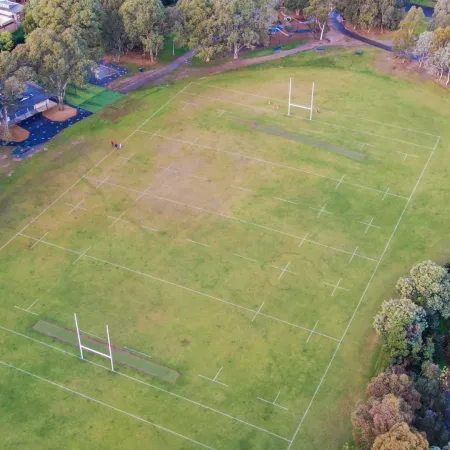 Aerial view of playground to the south and sportsground with rugby goal posts to the north. There are walking paths on each side of the playground and thick mature trees surround the area.