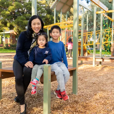One adult and 2 children sitting on a wooden structure in a playground. There is pine bark on the ground and trees in the distance.