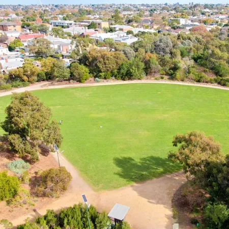 Aerial view of long, thin grass area with a straight walking path to the left, and a surrounding layer of trees and path in a U-shape.