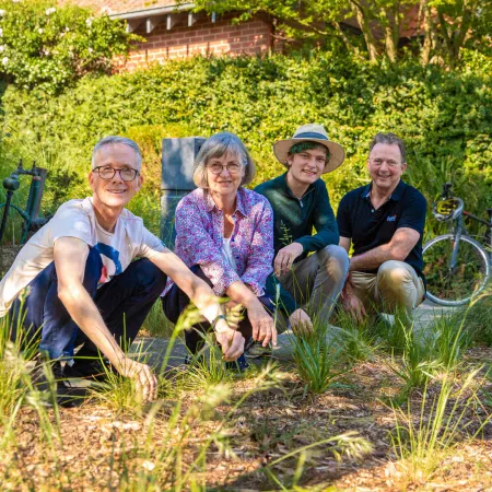 4 people crouching in a garden bed smiling at the camera. in the background is a hedge and a brick house