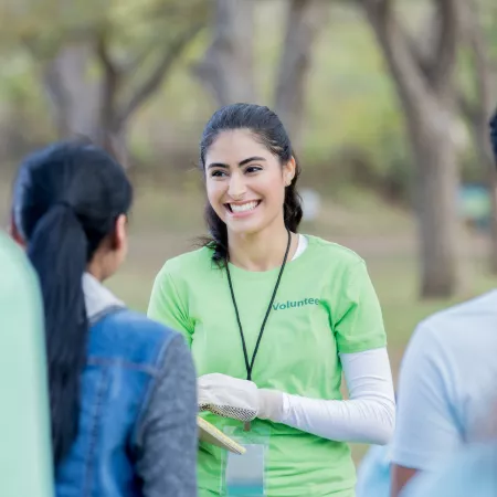 Young woman smiling with volunteer written on her t-shirt talking to group of people with backs to the camera
